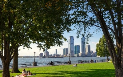 View of Jersey City skyline from a lush green park along the waterfront, with people leisurely relaxing on the grass.
