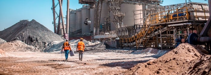Two workers in orange safety vests walk through a gritty industrial site with large silos and piles of materials in the background.