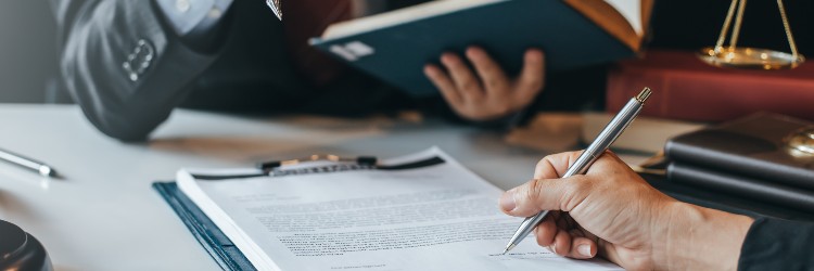 A close-up of a hand holding a silver pen, signing a document, while another person reads a book in a legal setting.