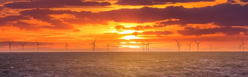 Vibrant sunset over the ocean with silhouetted wind turbines dotting the horizon, creating a serene and beautiful landscape.